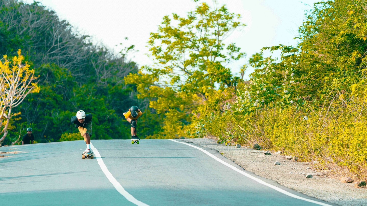 Several downhill skateboarders, including Anna Pixner wearing a yellow T-shirt and black helmet, are riding down a rural, slightly sloped road. The scene is surrounded by lush vegetation and trees.